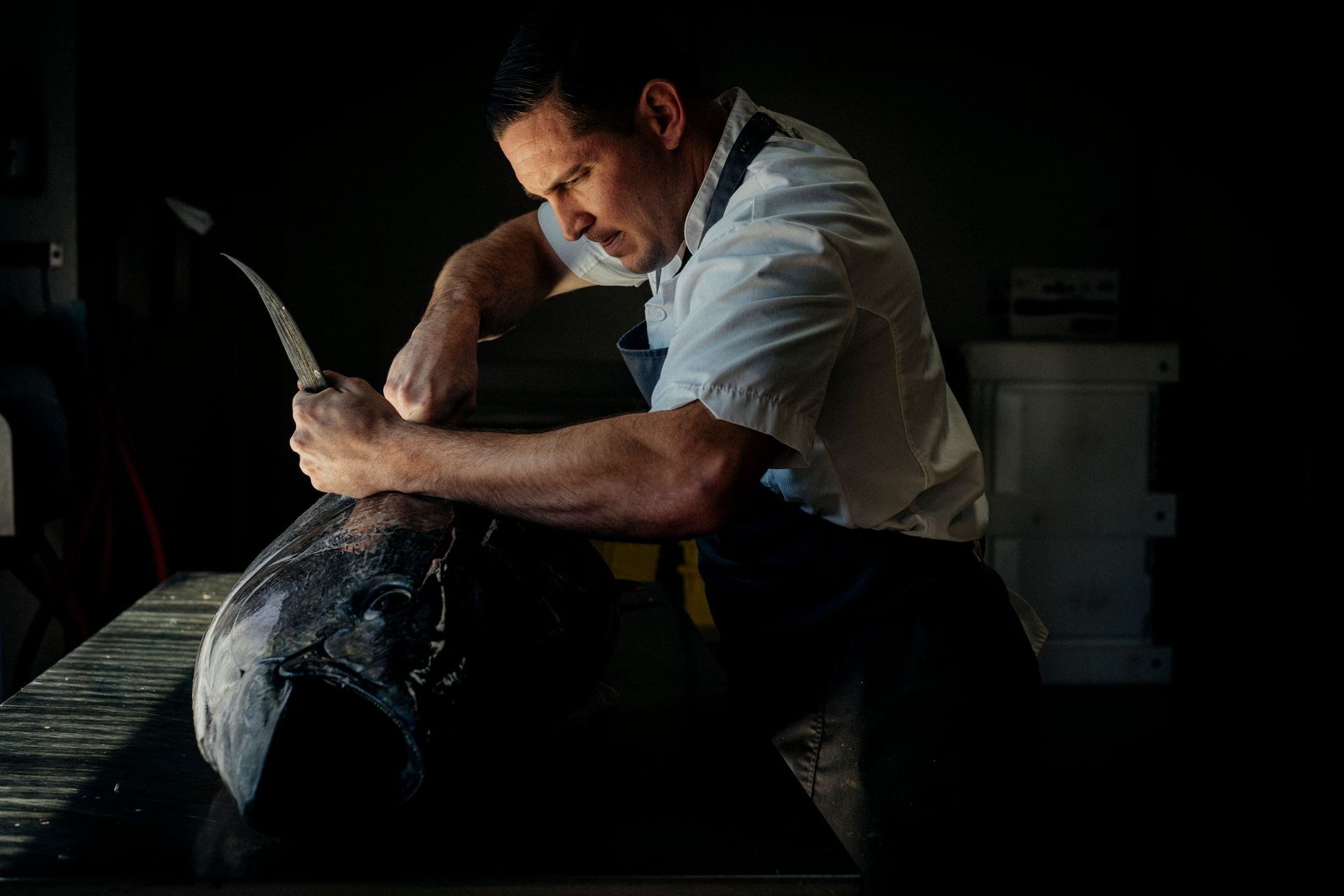 Chef in white apron filleting a large fish on a dark kitchen counter.